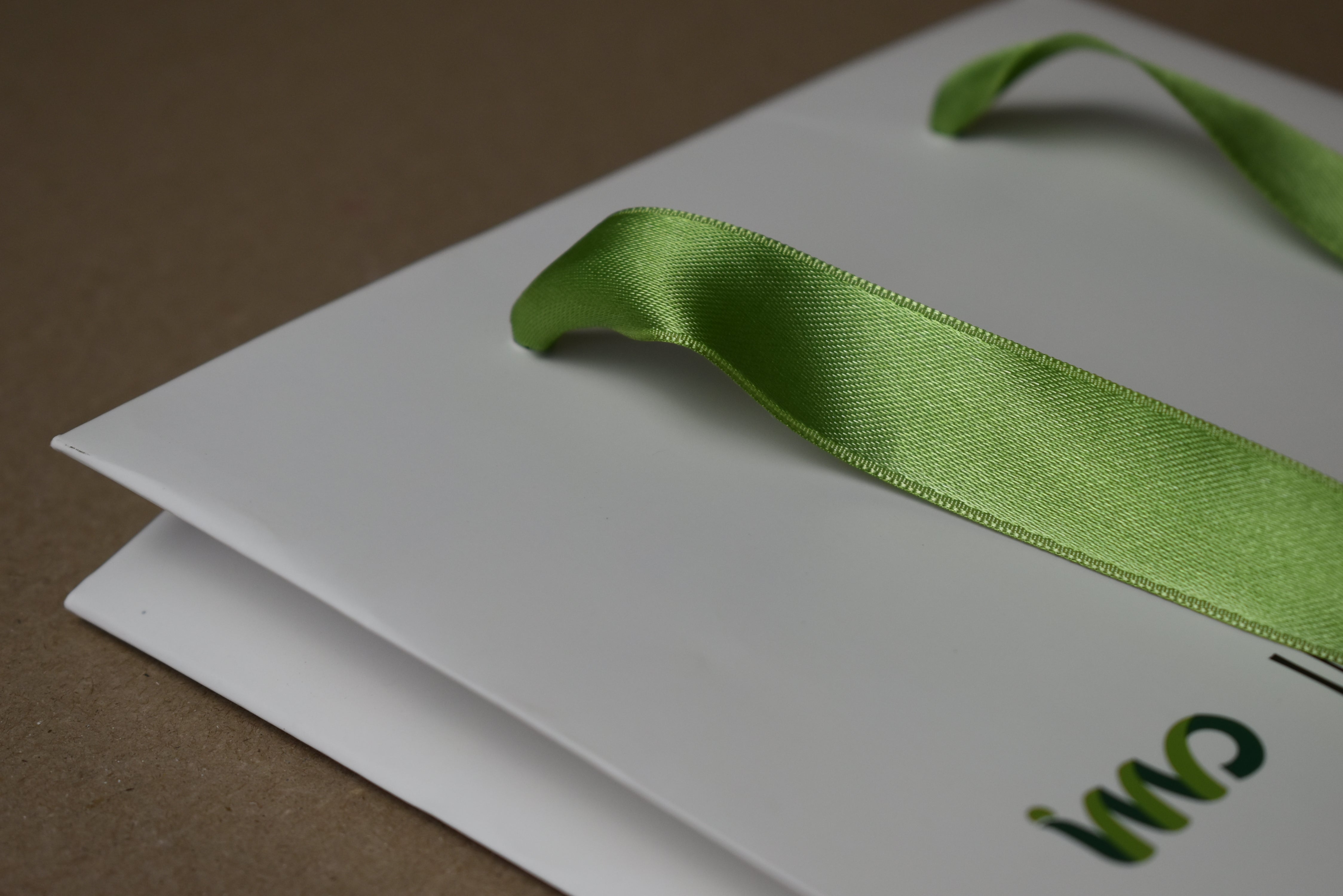 A close-up of a white paper gift bag with a light green satin ribbon handle, placed on a brown surface. Part of a green printed logo is visible on the bag.