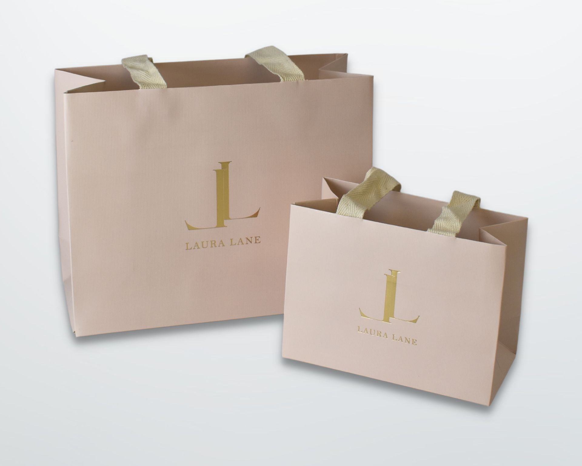 Two light blush pink paper shopping bags with gold foil 'LAURA LANE' logos and gold ribbon handles, shown on a clean white background.